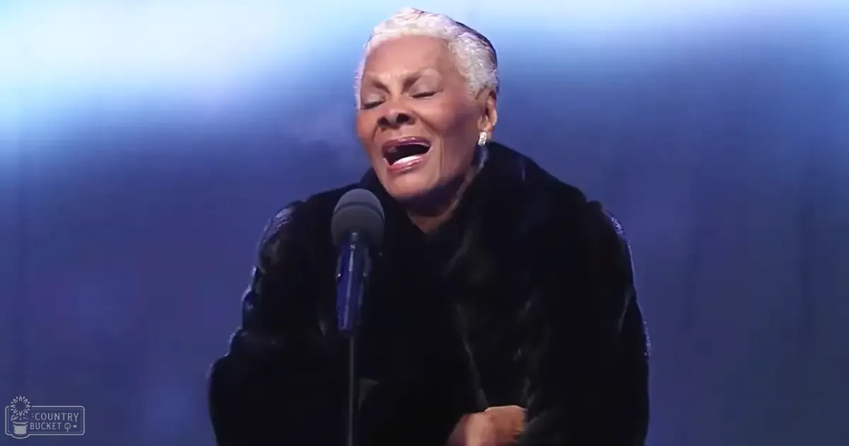 A middle-aged woman wearing a luxurious black fur coat performs on an indoor stage with blue lighting, holding a microphone.