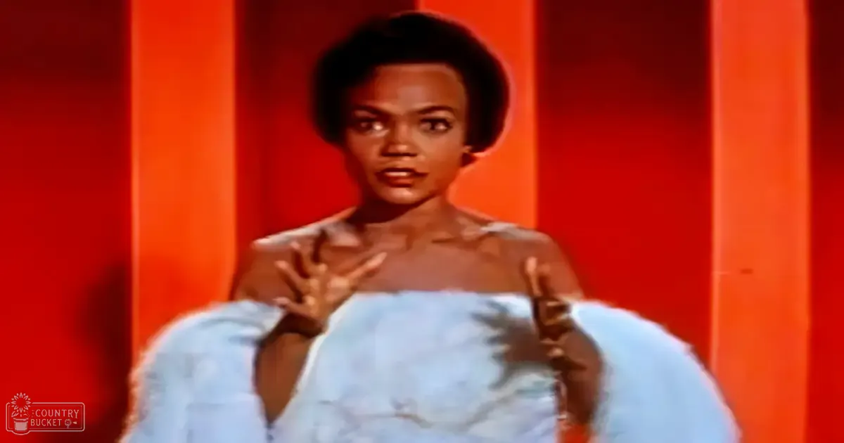 A young adult female in an indoor setting wears a light blue, textured garment resembling scales or leaves, likely mid-20th century, against vertical orange panels suggesting a theatrical backdrop.
