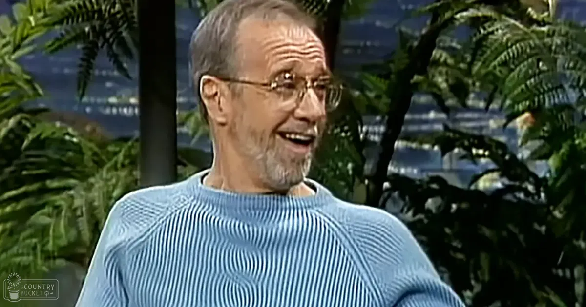 An older adult male wearing a light blue ribbed sweater in a brightly lit studio with large windows showing greenery and a cityscape view.