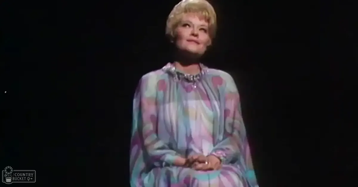 An older adult female in a performance setting, wearing a loose-fitting long-sleeved dress with an abstract floral pattern and bead necklace.