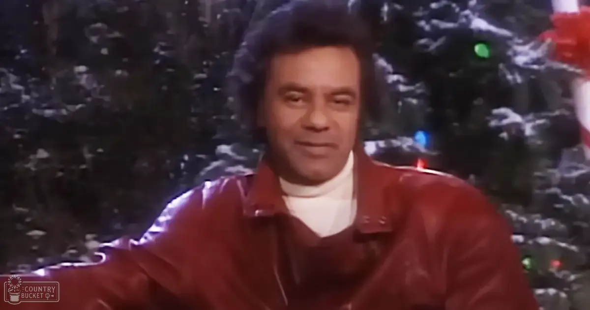 A middle-aged man wearing a red leather jacket and white turtleneck sweater stands against dark, textured walls with a Christmas tree decorated with lights in the background.