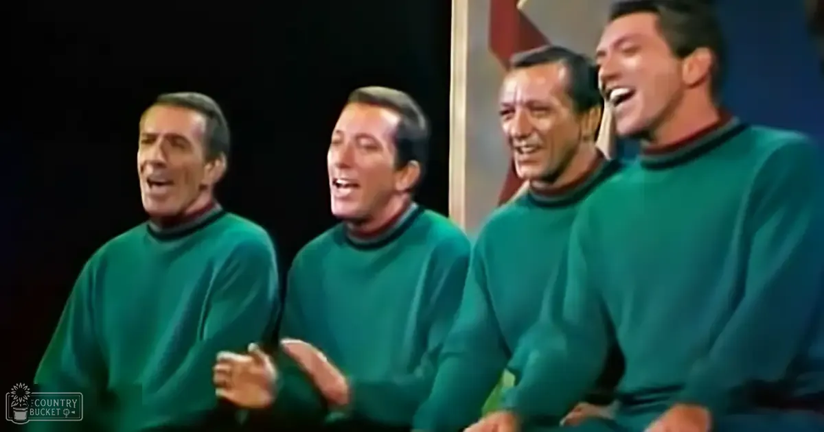 Four adult males singing together, wearing similar long-sleeved green sweaters with ribbed necklines and cuffs, indoors with controlled lighting for a performance or recording session.