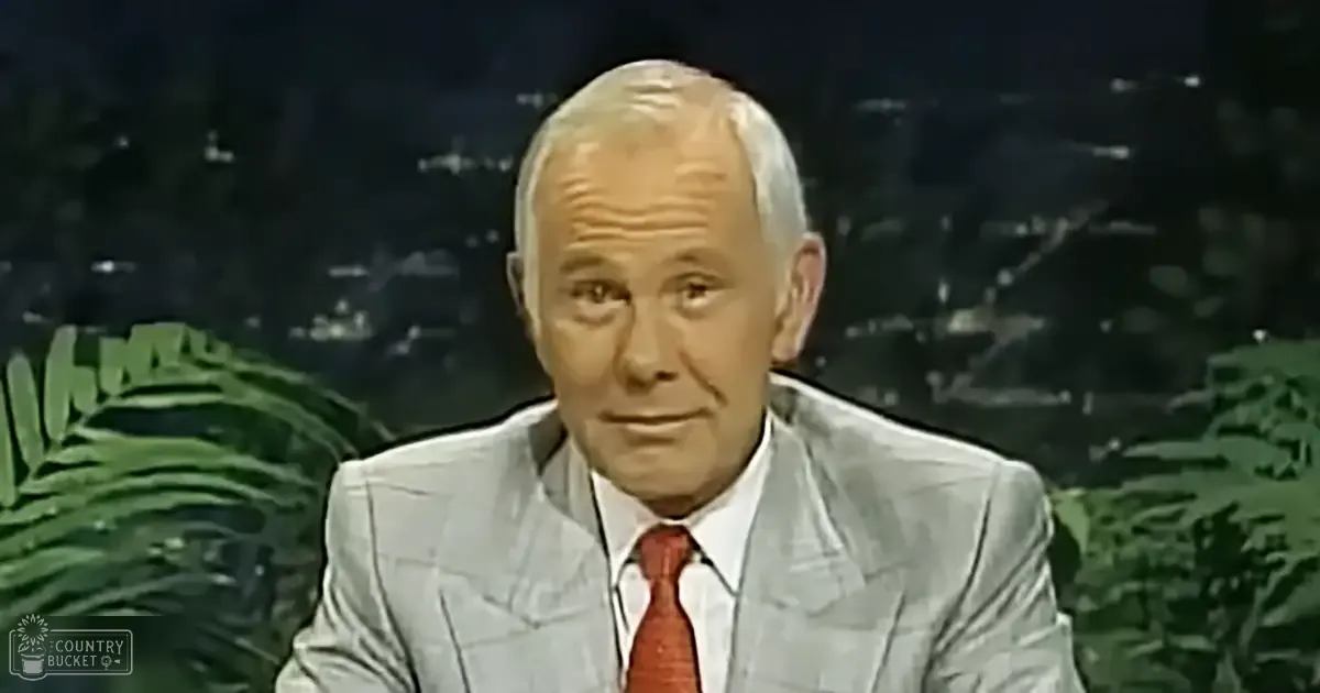 An older adult male in a mid-20th century style light gray pinstripe suit jacket, white dress shirt, and red tie against a dark backdrop with green foliage on either side.