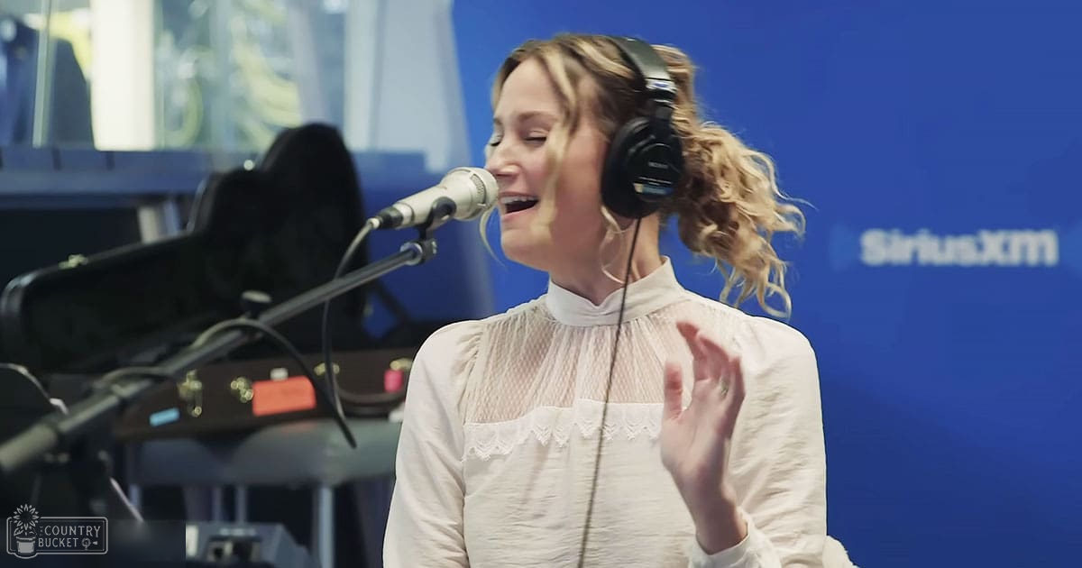A young adult female in a white blouse with ruffled neckline and black over-ear headphones stands in a recording studio setting, with SiriusXM signage visible on the blue wall.