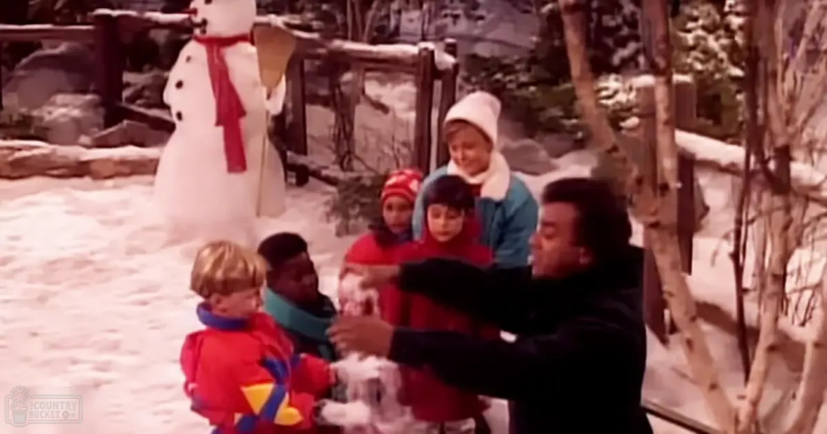 Children playing with an adult in a snowy outdoor setting, dressed warmly for winter; a snowman and wooden structures are part of the background.