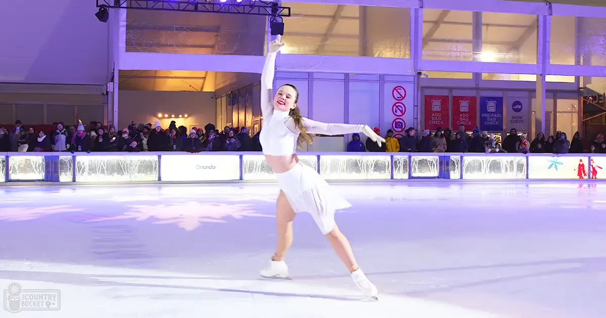 An indoor ice skating rink with a young female figure skater performing, surrounded by an audience in winter attire, and festive decorations indicating it might be during the holiday season.