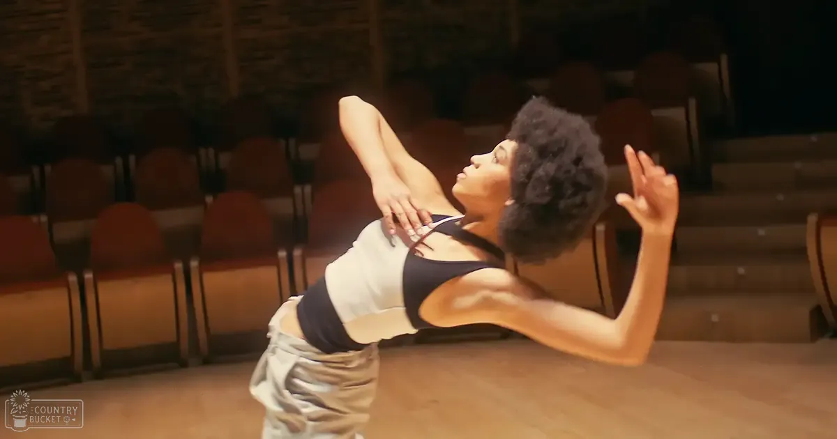 A young adult female dancer in an auditorium wearing a black and white sports bra with thin straps and light gray sweatpants, surrounded by rows of brown chairs under natural daylight.