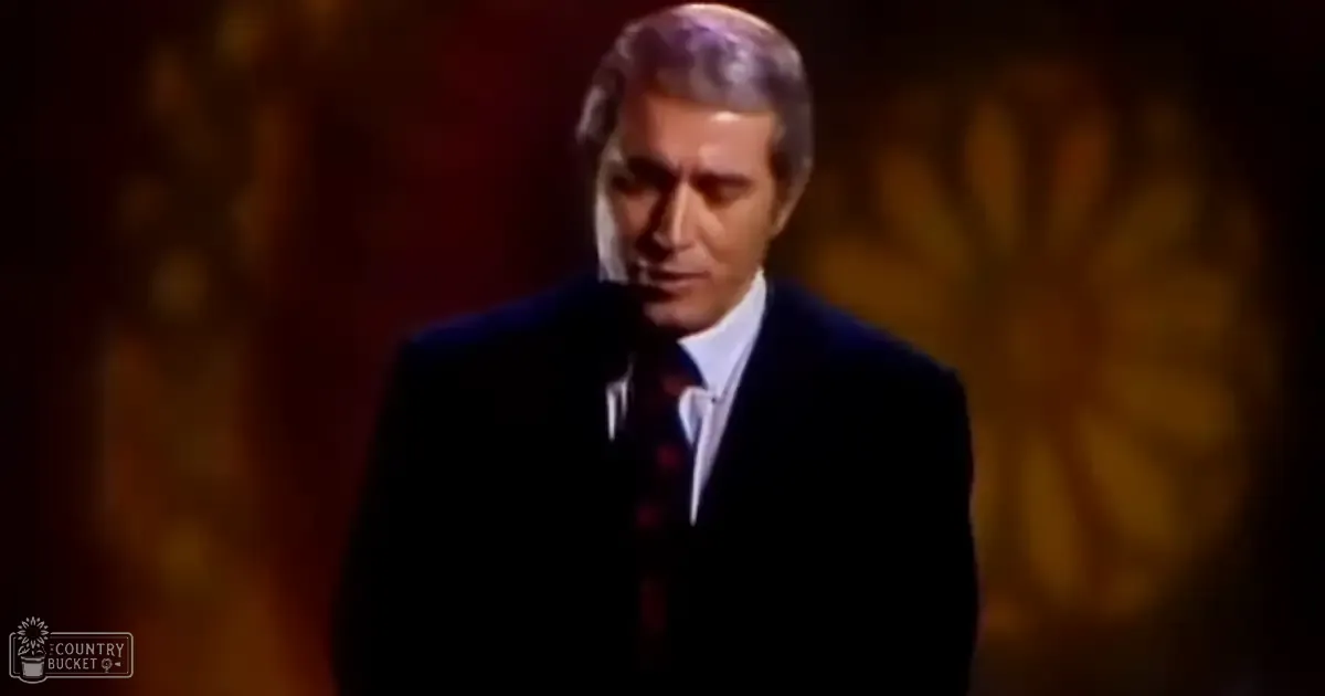A middle-aged man in a dark suit jacket and patterned red tie stands indoors against a warm-toned floral backdrop.