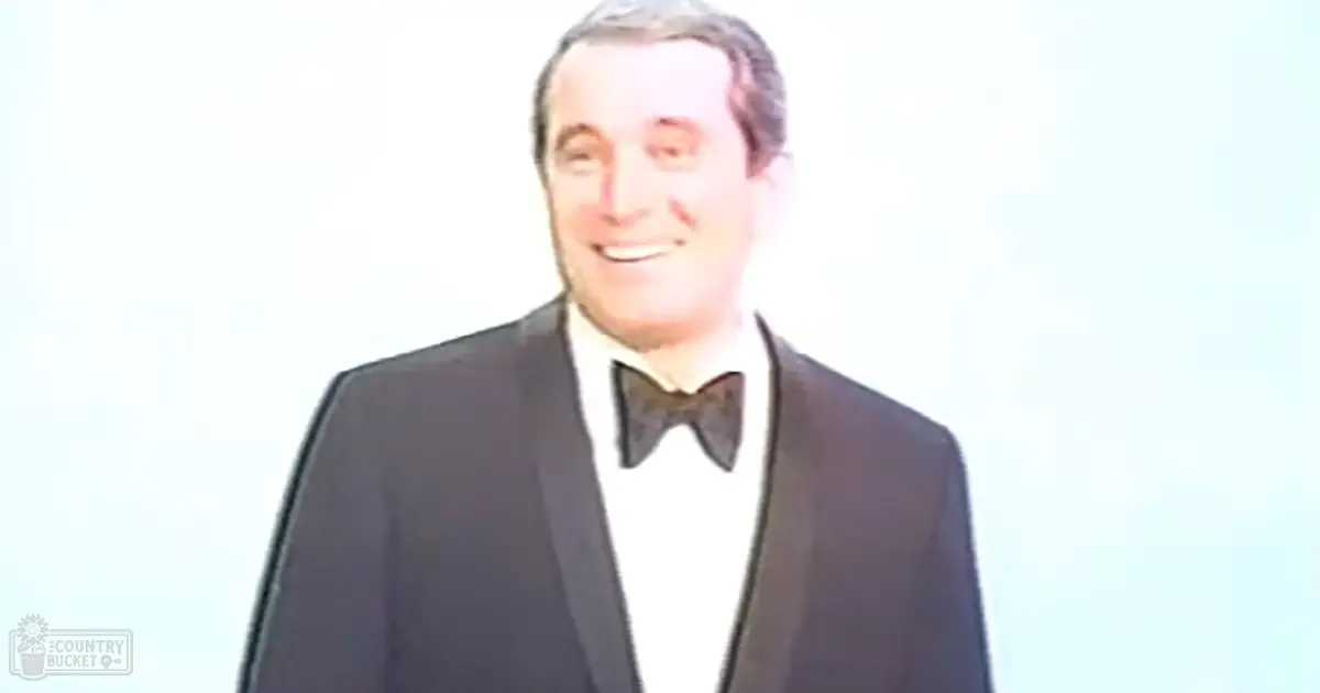 Adult male in a classic tuxedo with white shirt and black bow tie stands against a plain white background under bright daylight.