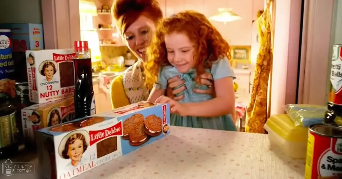 A young adult female wearing a yellow polka-dotted top and a toddler girl in a light blue dress are interacting in their home kitchen, surrounded by Little Debbie products on the table.