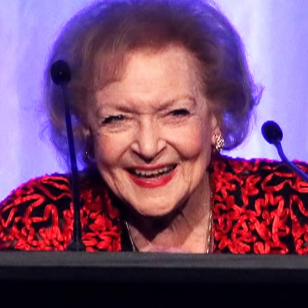 An older adult female in a red and black patterned dress with silk or satin material, wearing earrings and a necklace, positioned at what appears to be a formal event with microphones nearby.