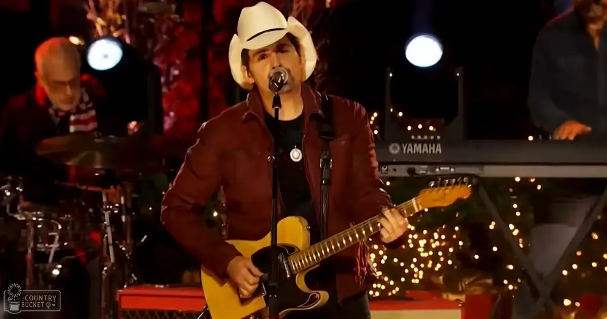 Adult male singer wearing a white cowboy hat with black band, maroon jacket, dark shirt, silver necklace, performing on stage at an indoor concert venue decorated for Christmas.