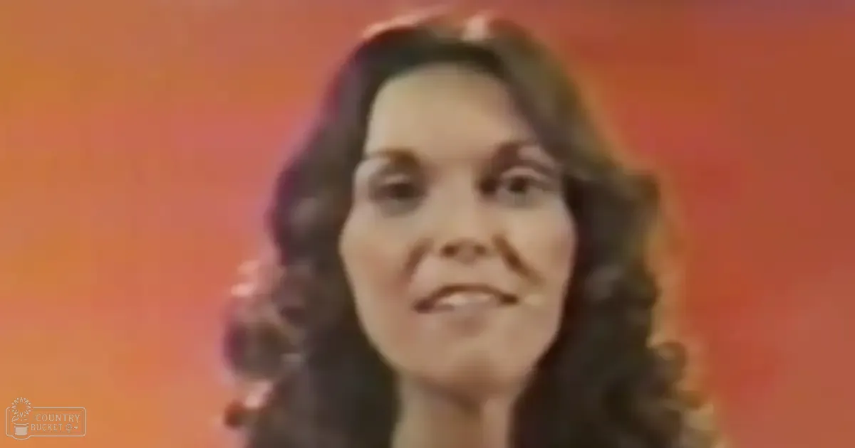 An adult female with long wavy dark hair indoors wearing a rounded neckline blouse or dress from the 1970s, set against an orange background.