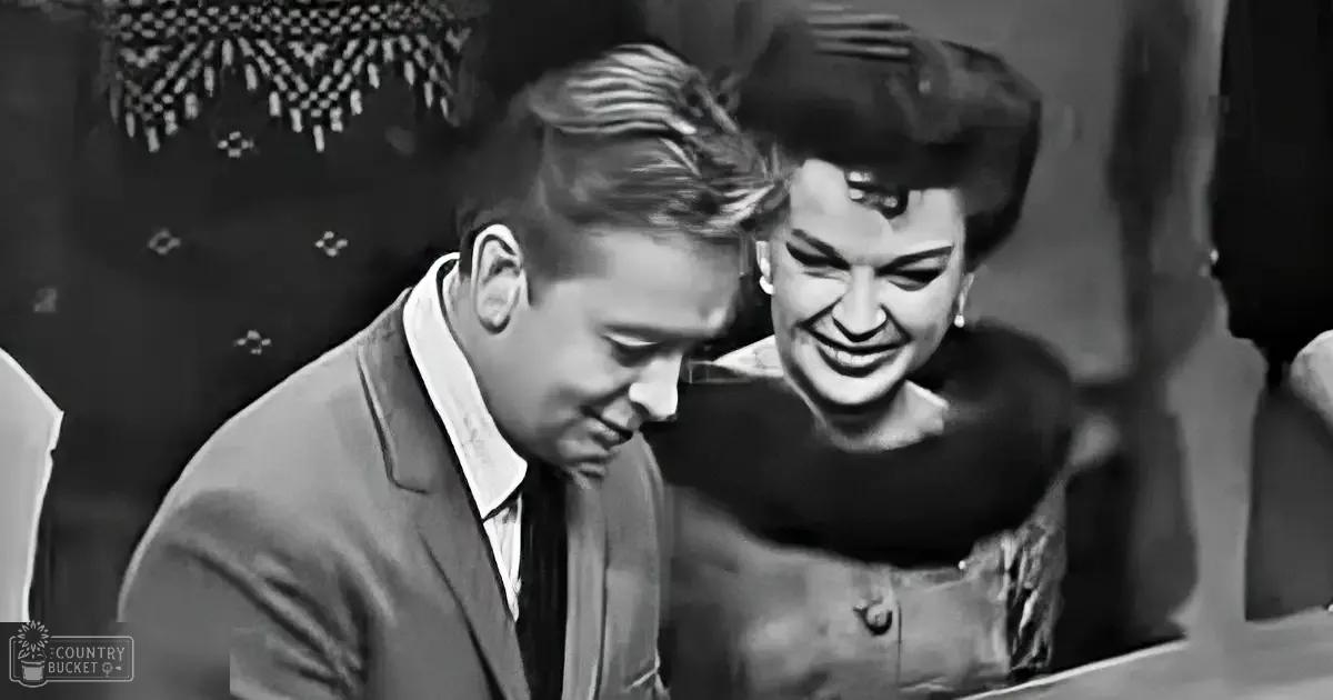 A young adult male in a dark suit jacket plays piano beside an older adult female in a mid-20th-century satin dress, both seated in what appears to be a studio or performance space with patterned backdrop and bright even lighting.