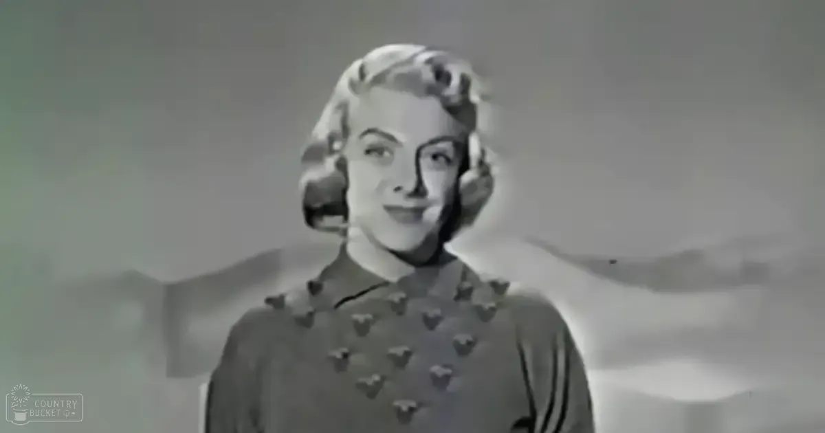 An adult female wearing a dark garment with small heart shapes stands indoors against a plain wall.
