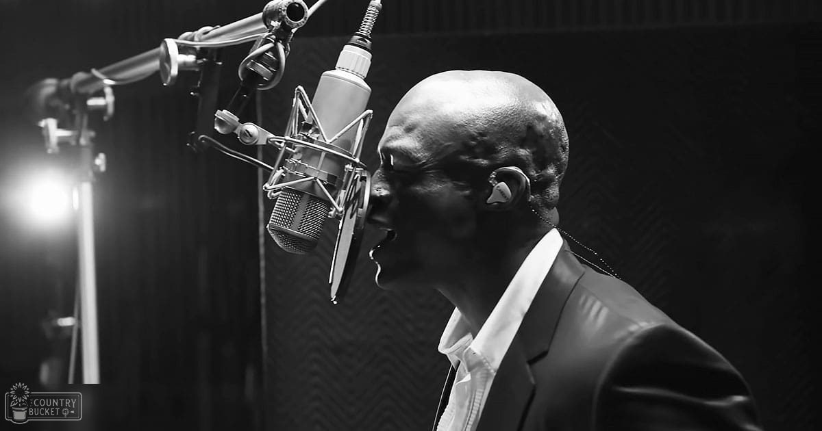 Middle-aged male adult wearing a dark suit jacket in a recording studio with microphones and soundproofing panels, illuminated by artificial light during evening hours.