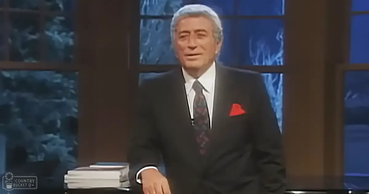 A middle-aged man wearing a dark suit with a red and black patterned tie in an indoor television studio setting.