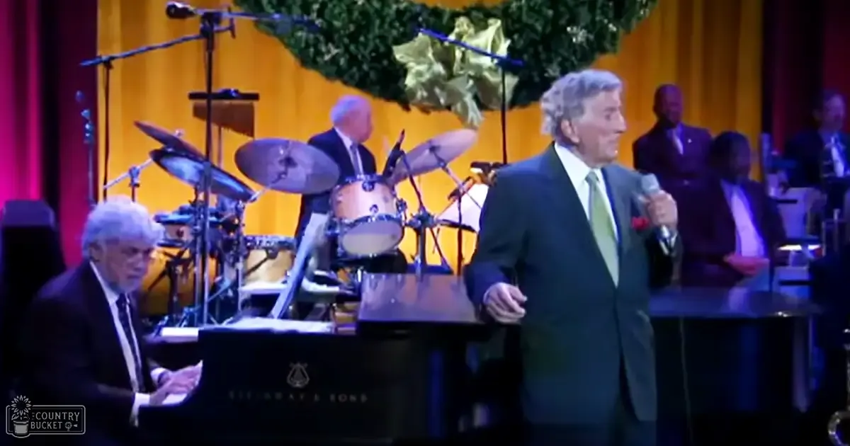 A male singer wearing a dark suit jacket, light green tie, and white shirt performs in front of an audience with a pianist in a formal concert setting. Microphones on stands and drums set up behind the pianist suggest a live musical performance.