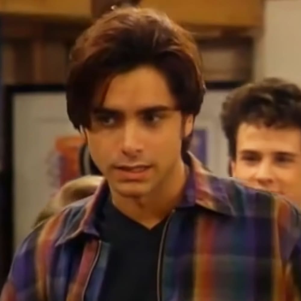 A young adult male wearing a plaid shirt with vertical stripes stands indoors, possibly in a home setting, during daytime.