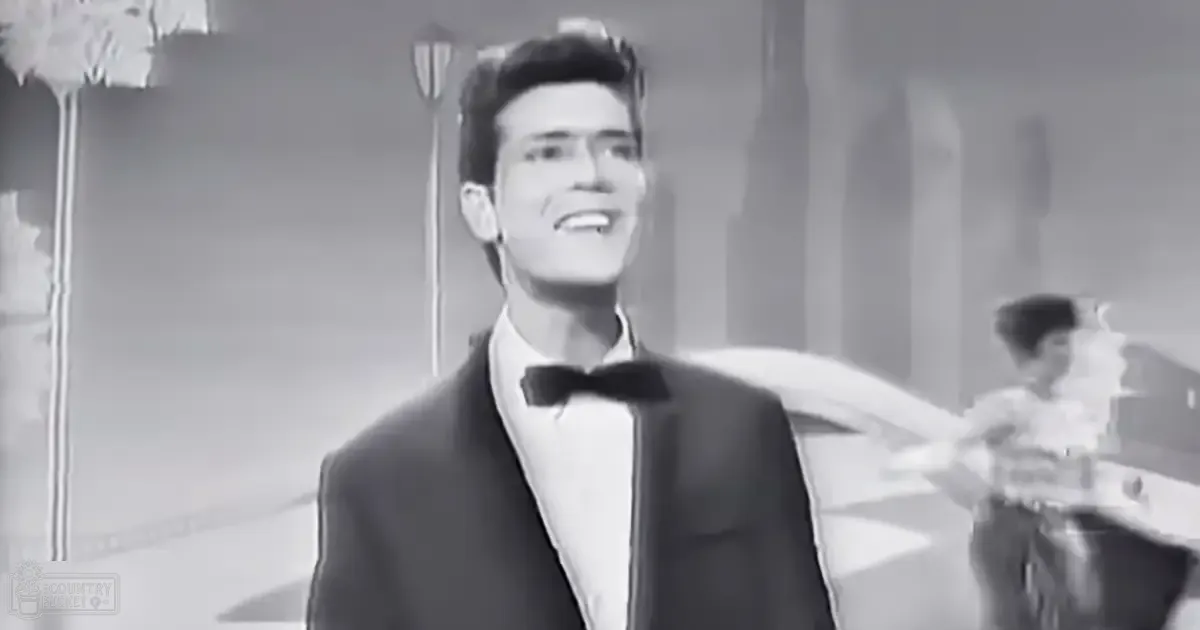 A young adult male in a formal black tuxedo stands on an indoor stage with curtains and decorative elements typical of mid-20th century productions.