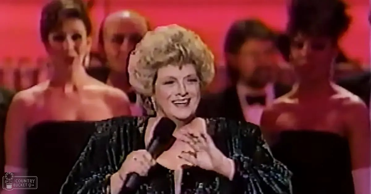 A middle-aged woman in a black, intricately detailed sequined 1980s-style dress speaks into a microphone at an awards ceremony with blurred figures of other attendees visible in the background.