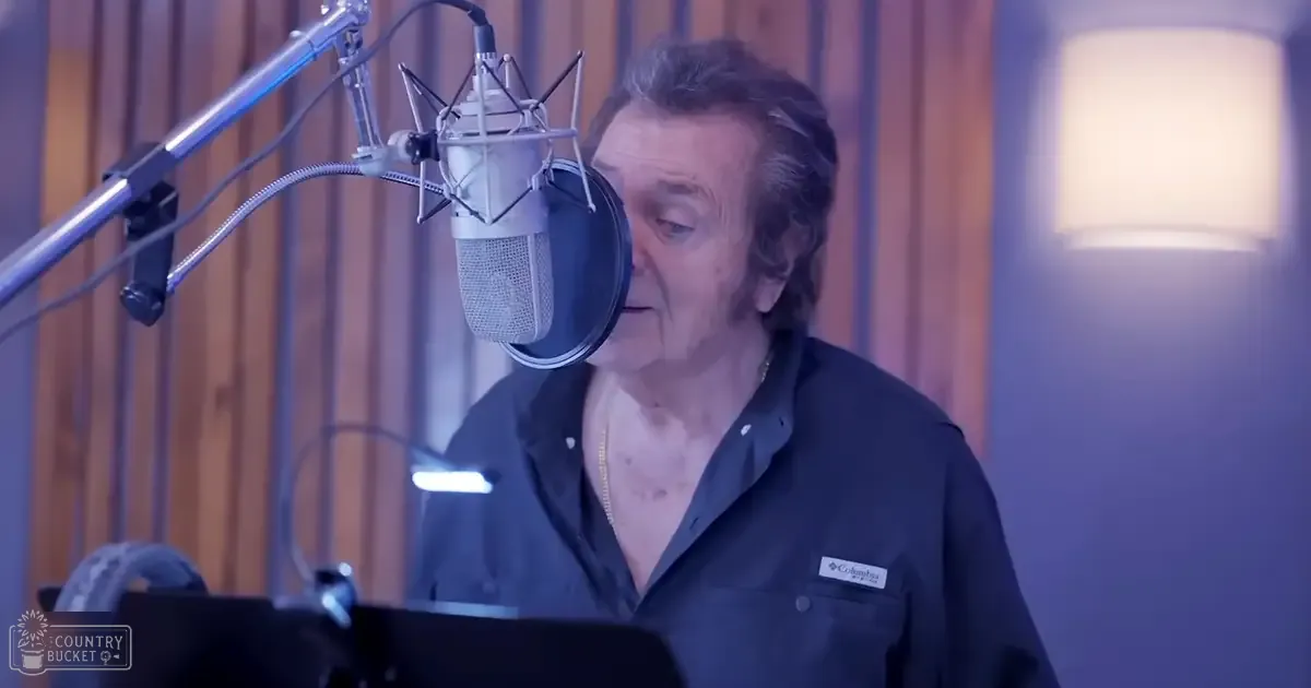 A middle-aged adult male in a recording studio, wearing a relaxed fit dark blue button-up shirt with a collar and chest pocket, holding sheet music or lyrics on a stand.