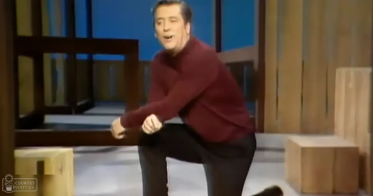 A middle-aged adult male in a maroon long-sleeved sweater stands indoors, possibly at a performance venue with blue lighting.