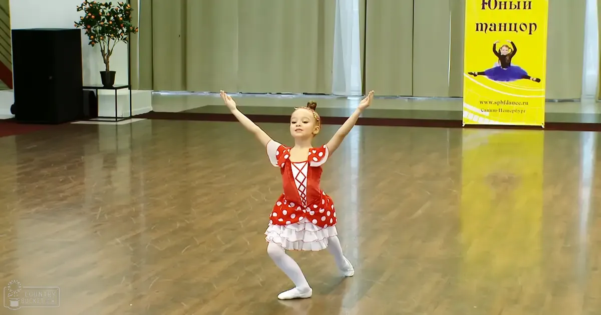 A child in a dance studio wearing a red and white polka-dot ballet dress with ruffled skirt, white ballet slippers, and a small headband with a bow.