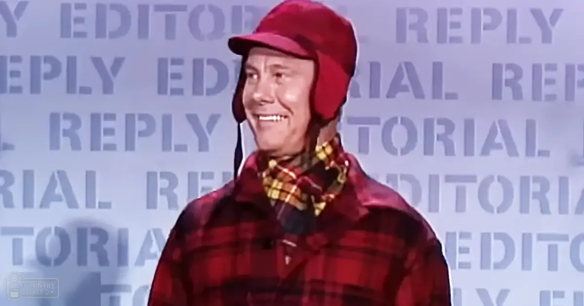 A middle-aged adult male wears a red plaid jacket with a high collar and button closure, set against a backdrop with 'EDITORIAL REPLY' text.