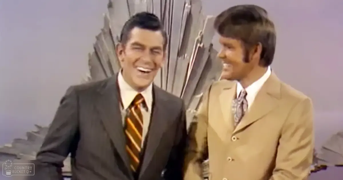 Two adult male singers wearing classic tailored suits, one with a dark gray suit and orange striped tie, the other with a beige suit and patterned tie, set against a studio backdrop for what appears to be a talk show or similar program.