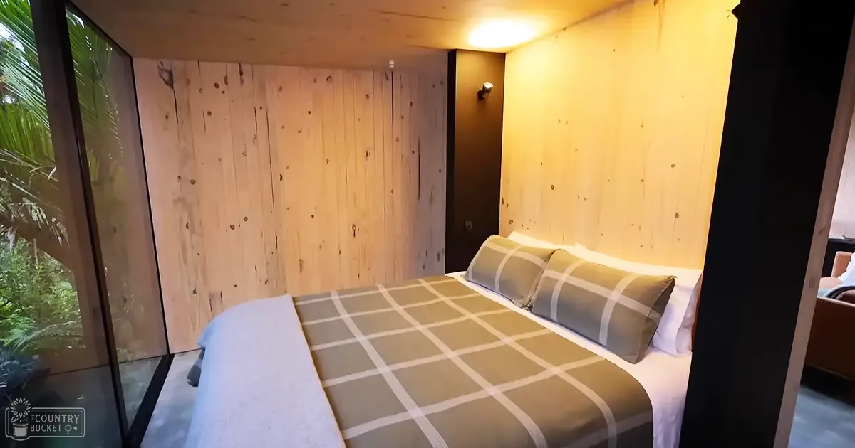 A modern bedroom with a wood-paneled wall, a bed with white sheets and a green checkered blanket, two pillows, and natural light streaming in from a sliding glass door leading outside.