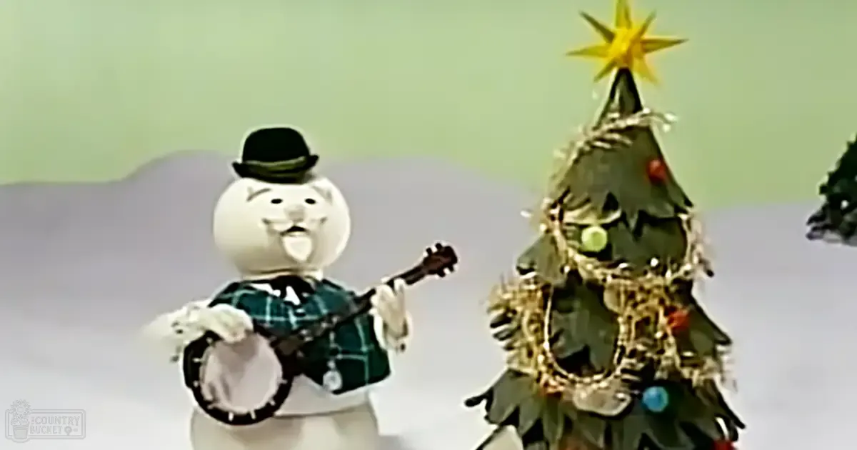 A snowman playing a banjo next to a decorated Christmas tree indoors, with white, black, green, red, gold, and yellow dominant colors.
