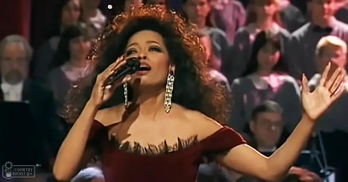 A female performer singing on stage at a formal event, wearing a strapless red dress with feather-like details and large dangling earrings.