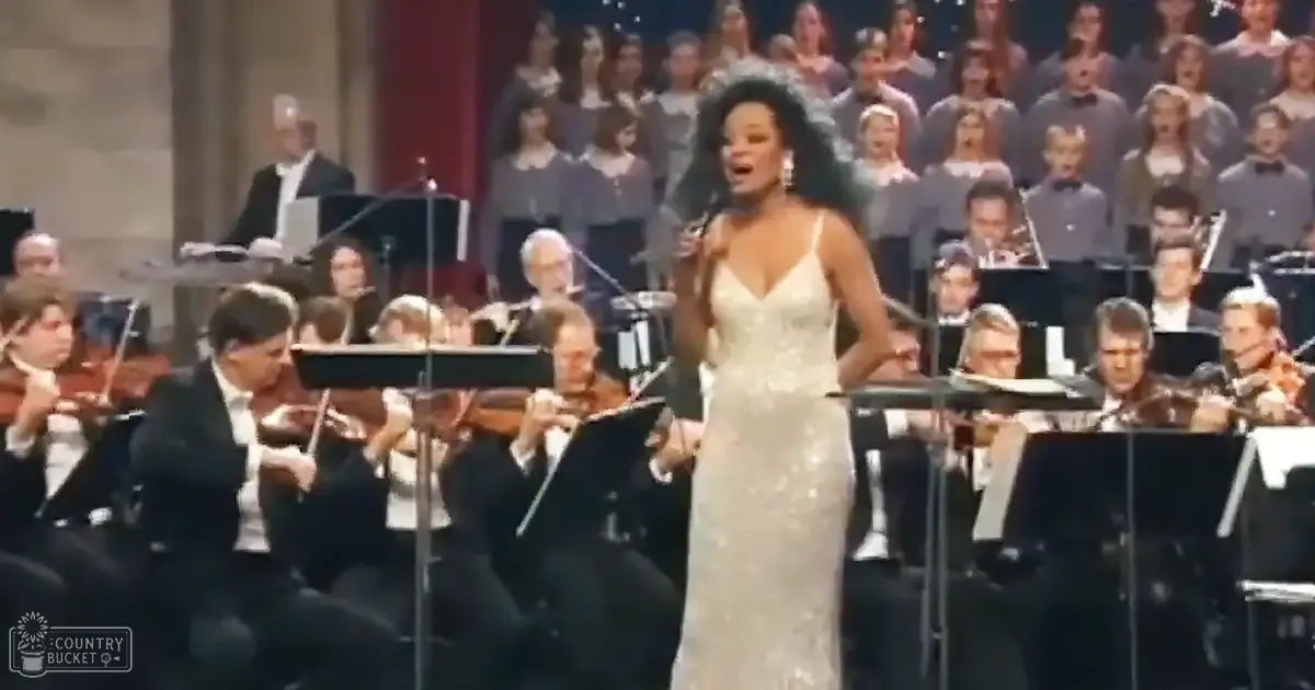 A woman in a white, sequined 1930s gown sings on stage at a concert hall with an orchestra and choir.