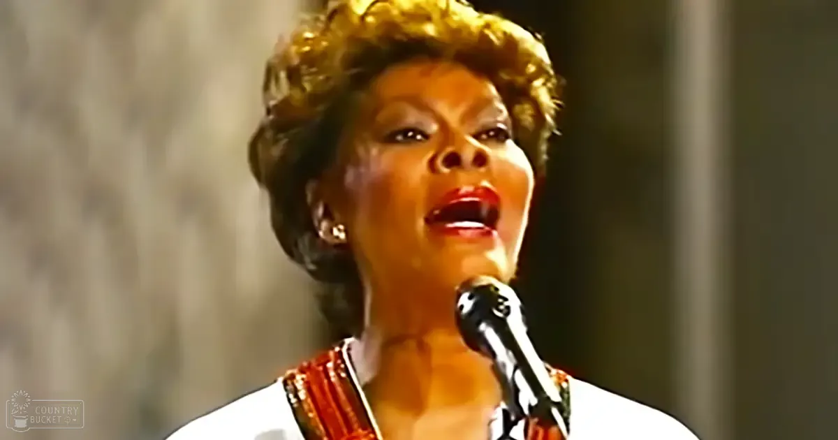 A woman singing into a microphone on an indoor stage, wearing a white garment with red accents in mid-20th century formal attire.