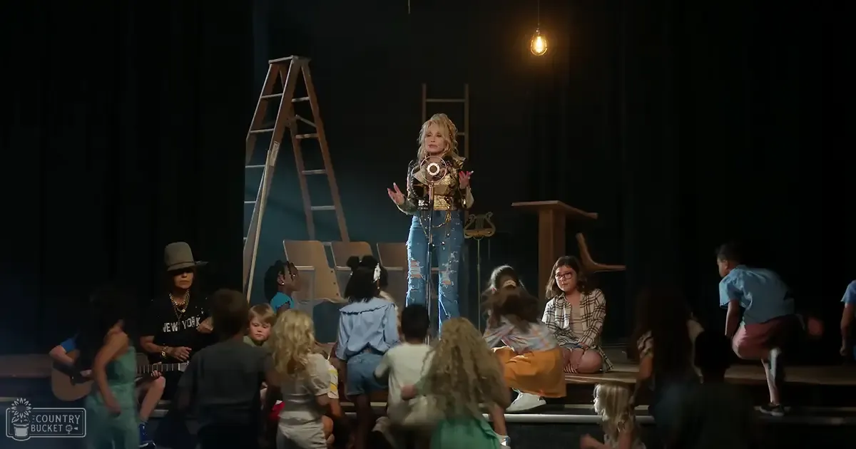 A middle-aged female performer in a theater setting, surrounded by an audience of children wearing casual clothes under stage lighting at night.