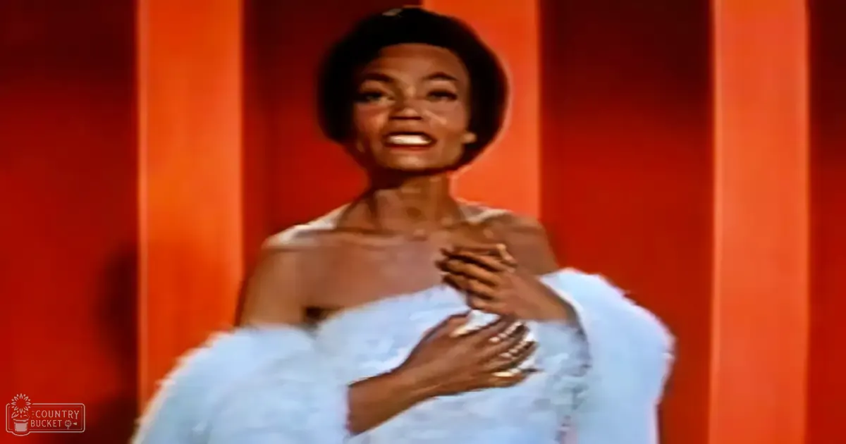 An adult female in an indoor setting with a light blue, fluffy shawl draped over one arm against a vibrant orange backdrop.