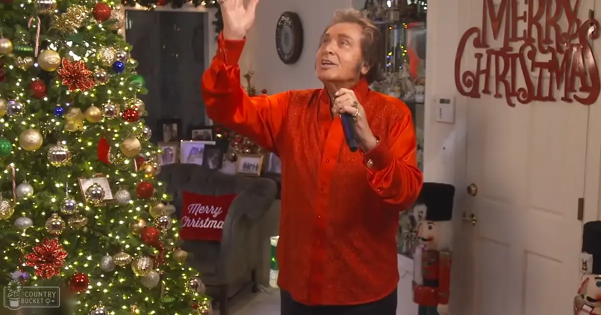 A middle-aged adult male in a relaxed, long-sleeved shirt with a red brocade-like pattern, holding a microphone and raising his arm, standing indoors decorated for Christmas.