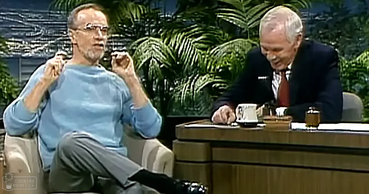 Adult male talk show host in studio with older adult male guest seated behind a desk filled with various items. Indoor setting with controlled lighting, spring or summer season suggested by greenery.