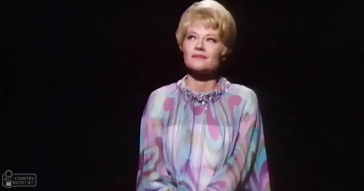 A middle-aged female wears a loose, ruffled, pastel floral blouse under stage lighting with a spotlight focused on her, set against a dark background.