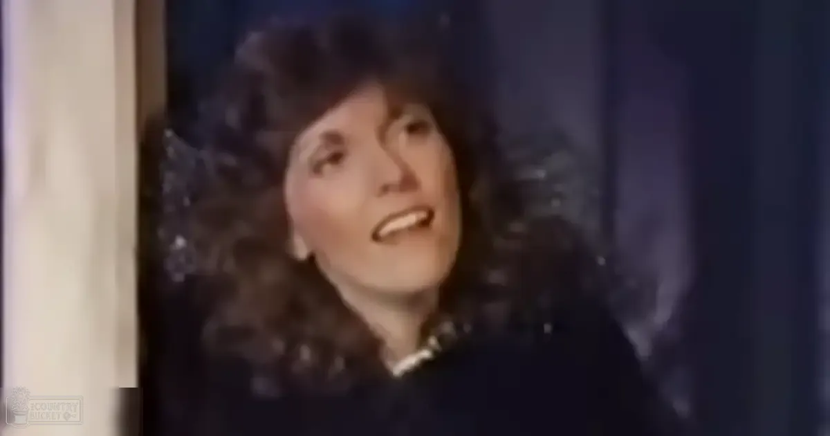A middle-aged adult female dressed in a dark-colored formal garment with a high collar stands indoors under dramatic lighting against a backdrop of dark curtains or a studio environment.