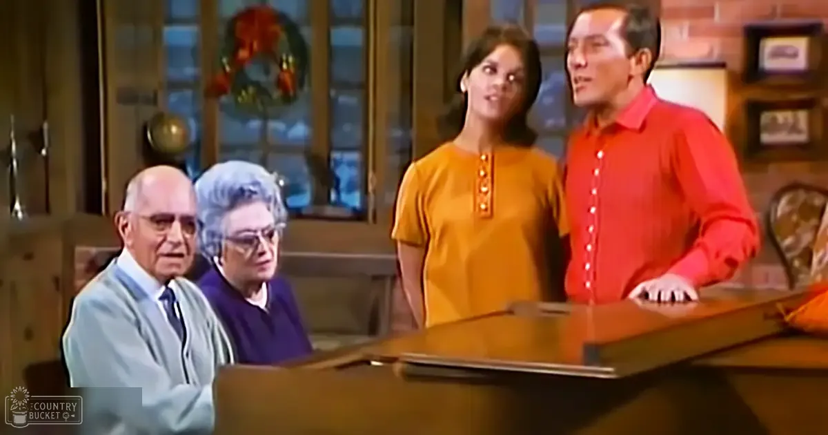 A family scene of four adults gathered indoors around a piano, with wooden furniture and Christmas decorations in the background.