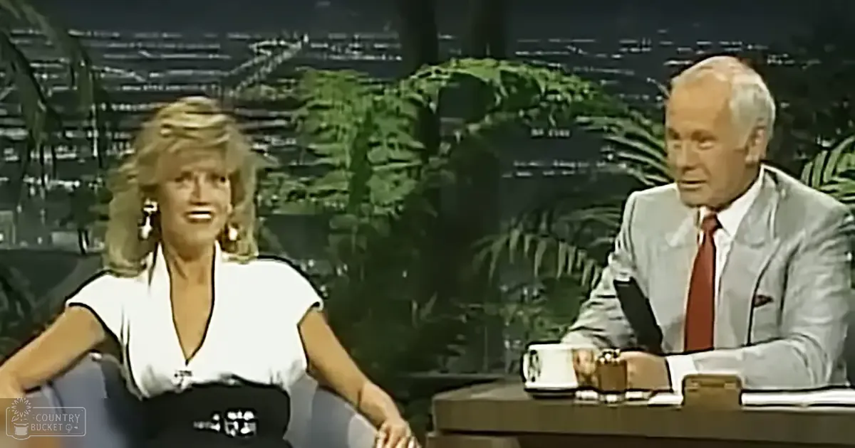 Talk show host and guest in an indoor studio setting with lush greenery and a cityscape backdrop.