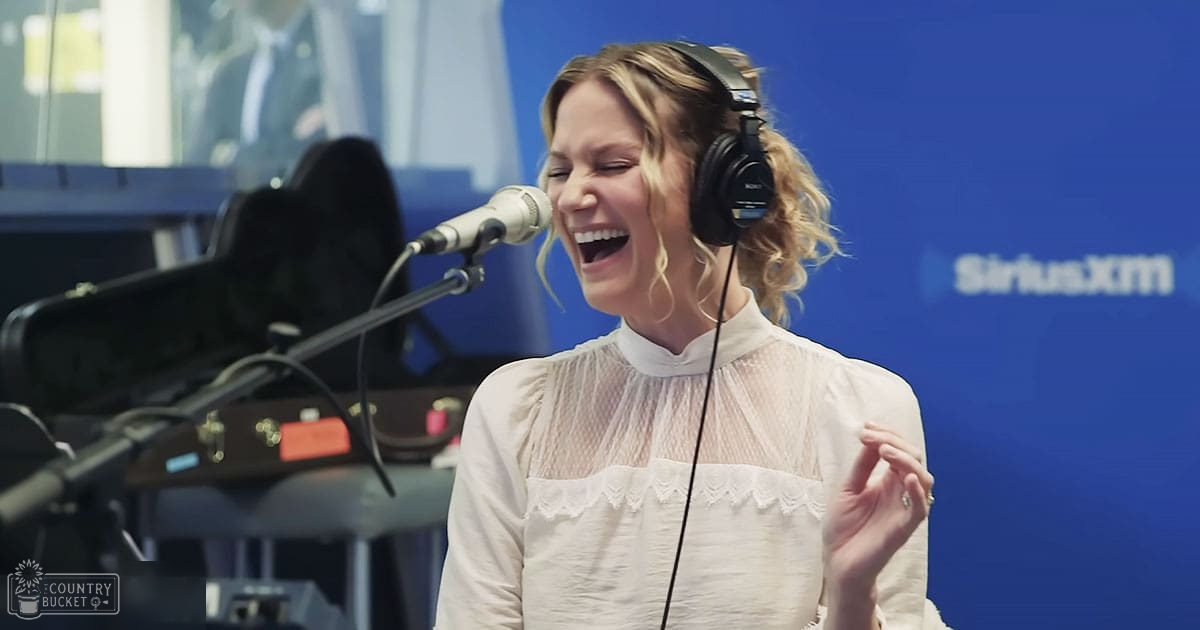 A young adult female in a recording studio at SiriusXM, wearing a white lace blouse with ruffled edges in front of a blue backdrop.
