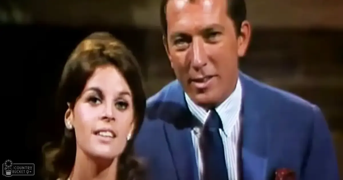 A middle-aged man in a blue suit jacket stands beside a young adult woman wearing a dark dress with large hoop earrings, against an indoor backdrop that suggests a stage or studio environment.