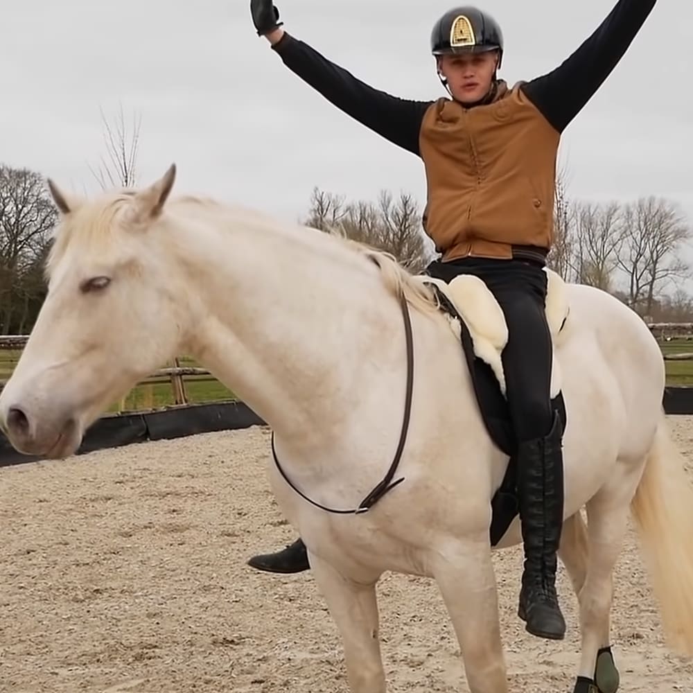 A young adult male in a black helmet with gold emblem, riding a white Arabian horse in an overcast outdoor equestrian arena surrounded by leafless trees suggesting autumn or winter.