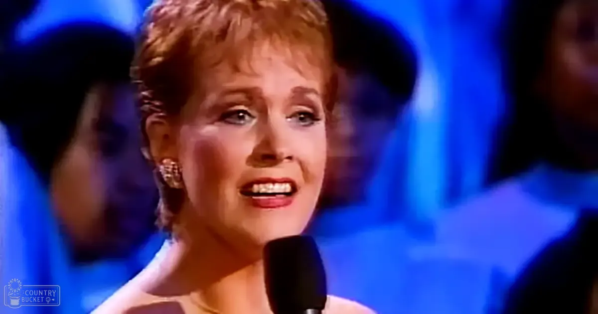 A middle-aged female performer singing into a microphone at an event or performance setting with stage lighting predominantly in blue and white, creating a dramatic effect.
