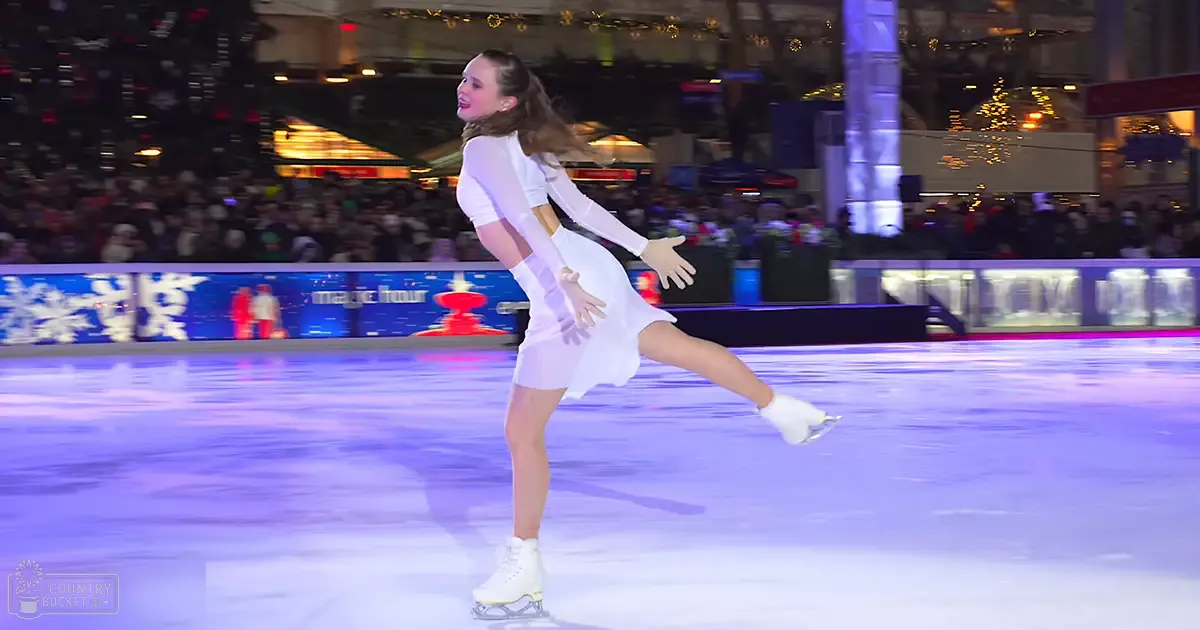 A young adult female figure skater performs at an indoor ice rink decorated for Christmas, with festive lights and holiday decorations visible in the background.