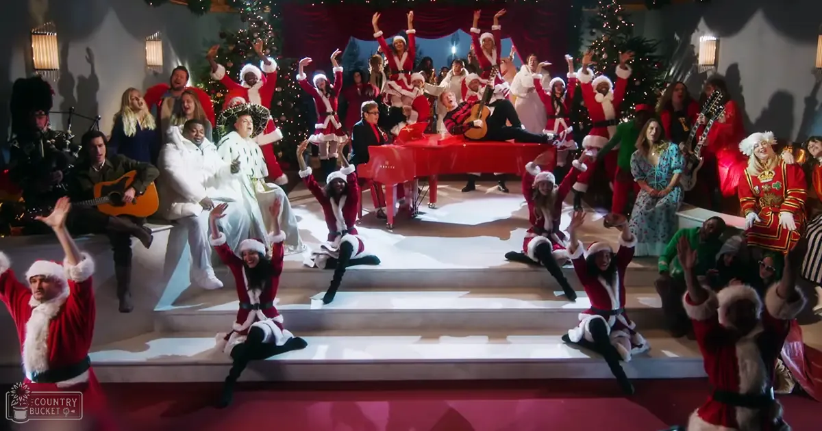 A festive Christmas performance scene with young adults dressed as Santa's helpers against an indoor studio backdrop adorned with decorated Christmas trees and bright lights.