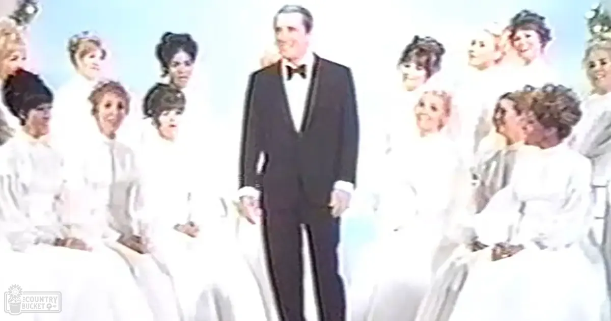 An adult male singer stands at center stage, dressed in a formal tuxedo with a bow tie and white dress shirt; two rows of women in light dresses are seated to the side against a plain white backdrop.